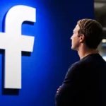 Mark Zuckerberg de espaldas contemplando el icónico logotipo de la letra F de Facebook sobre una pared azul iluminada.