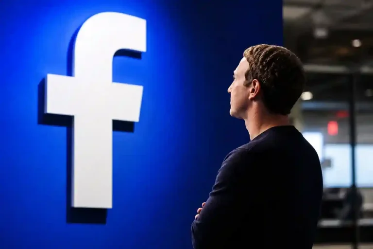 Mark Zuckerberg de espaldas contemplando el icónico logotipo de la letra F de Facebook sobre una pared azul iluminada.