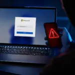 Portátil con pantalla de login de Microsoft y smartphone con icono de alerta roja, ilustrando el phishing de consentimiento en Microsoft 365.
