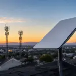 Antena Starlink sobre el techo de una vivienda con torres de telefonía celular tradicional al fondo durante el atardecer, simbolizando la competencia con AT&T, Verizon y T-Mobile